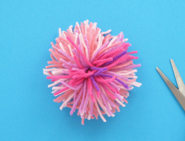 Ein Pompon aus Wolle in verschiedenen Rosatönen, mit einer Schere an der Seite.