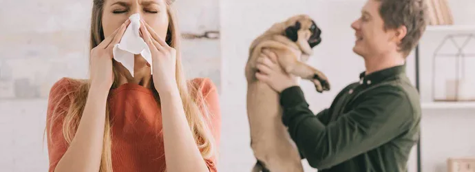 Μια γυναίκα φτερνίζεται την ώρα που ένας άντρας πίσω της κρατά στα χέρια του ένα σκυλάκι παγκ