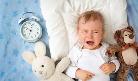 Ένα μωρό πάνω σε μια κουβέρτα κλαίει ενώ περιβάλλεται από δύο λούτρινα παιχνίδια και ένα αναλογικό ξυπνητήρι