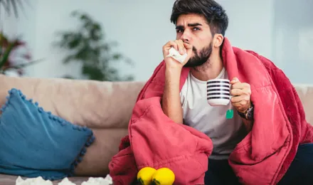 Молодой человек кашляет на диване под одеялом с кружкой чая и салфетками в руке