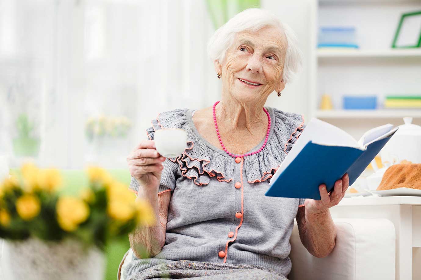 Пожилая женщина сидит на диване, держа книгу и чашку