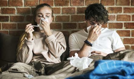 Две молодых людей сидят под одеялами и чихают в салфетки с фоном кирпичной стены