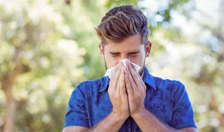 Un tânăr într-o cămașă albastră cu un fundal de pădure neclar strănută și se confruntă cu unele simptome ale alergiei la polen