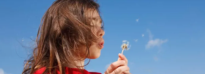 Malo dijete koje će možda trebati kućne lijekove za peludnu groznicu puše u maslačak s plavim nebom u pozadini
