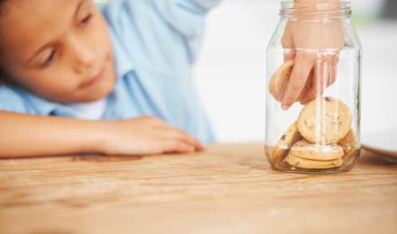 Ребенок дотягивается до стеклянной банки с печеньем на деревянном столе и берет печенье