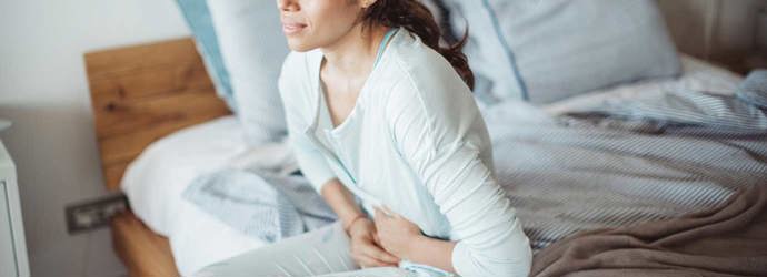 Eine Frau sitzt mit Schmerzen im Unterleib auf einem Bett