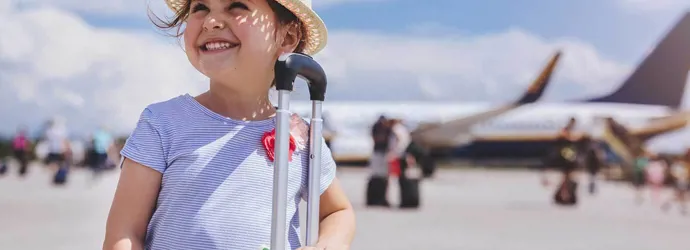 Ένα κορίτσι με αεροπλάνο στο φόντο χαμογελά ενώ κρατά μια κίτρινη βαλίτσα με τα απαραίτητα των οικογενειακών διακοπών