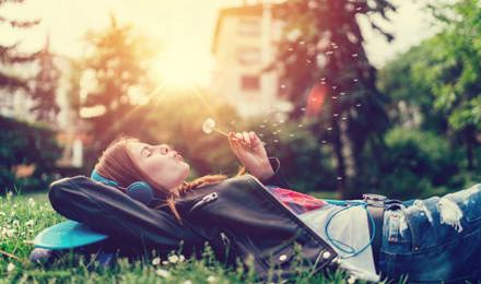 Eine junge Frau liegt auf dem Rasen, hört Musik und pustet eine Pusteblume